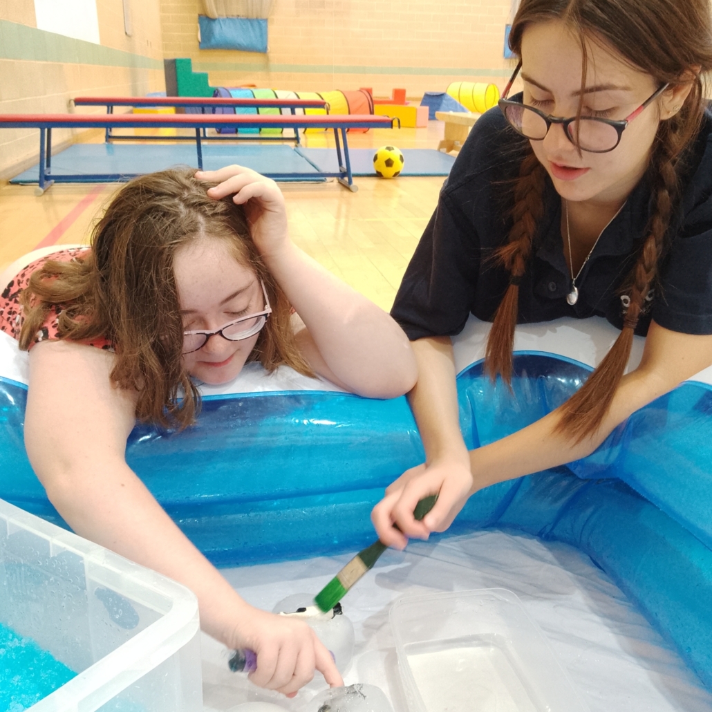 Accuro staff member with a Saturday Play Club member, excavating toys from ice with a paintbrush, in a paddling ppol